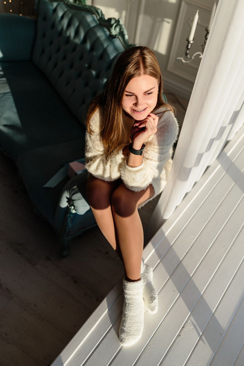 A young girl sits comfortably on a modern couch, smiling warmly as sunlight filters through sheer curtains, creating a cozy atmosphere in the stylish space.