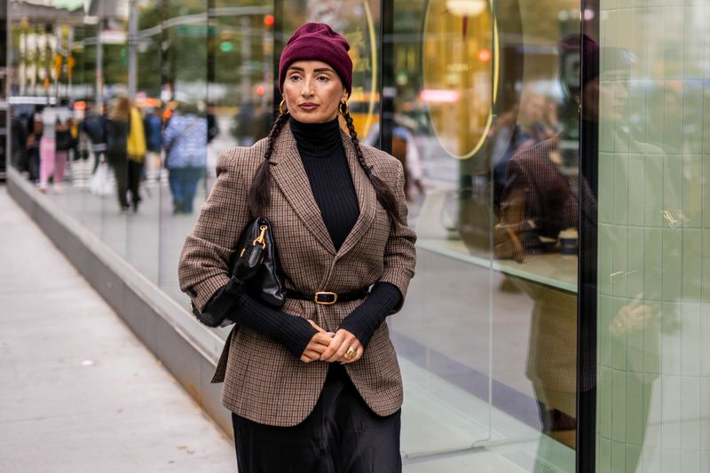 Fashionable brunette woman with braided hair in a burgundy cap, plaid jacket, black turtleneck and skirt, carrying a bag as she walks past a glass building in Manhattan, her reflection mirrored in the glass amid autumn city reflections.