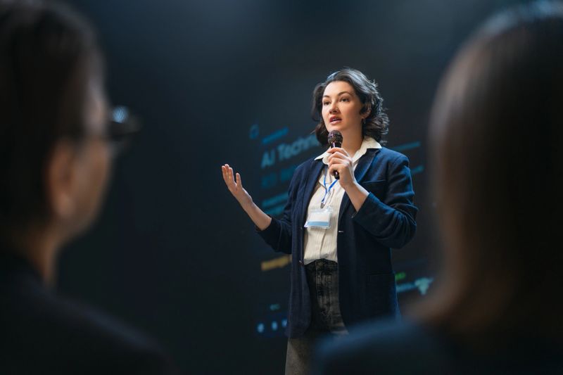An empowered businesswoman addresses an audience at a modern conference, holding a microphone and engaging listeners with her confident delivery. This image captures leadership, innovation, and professional communication, ideal for illustrating success, entrepreneurship, or public speaking in dynamic business environments.