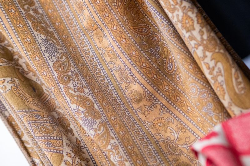 A high-detail close-up shot of a folded textile with a traditional paisley and floral motif. The fabric is arranged in soft diagonal drapes, highlighting the intricate print of ochre, mustard, and earthy brown stripes separated by delicate white and purple geometric borders