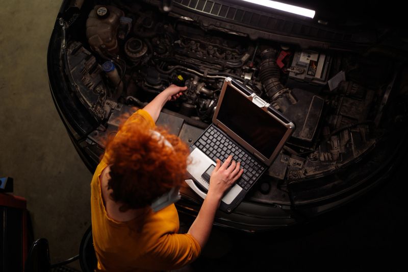 Young adult woman technician working on a car engine, performing a diagnostic check with a laptop in a dimly lit garage, representing modern vehicle service and maintenance