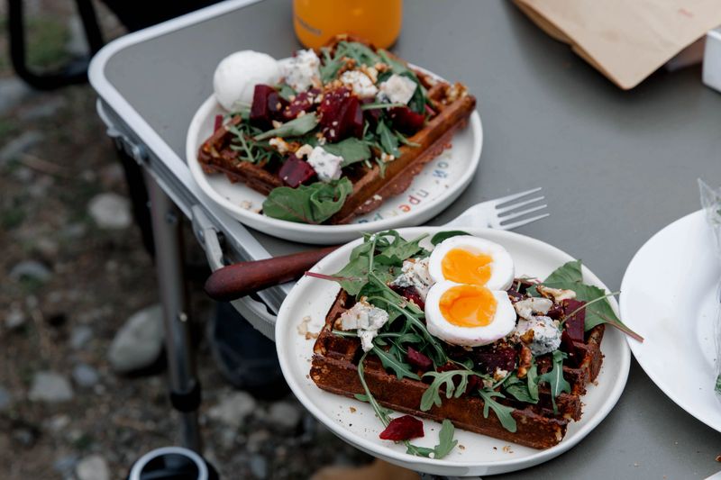 Two plates of gourmet waffles topped with greens, beets, and soft-boiled eggs. A refreshing drink is nearby on a table.
