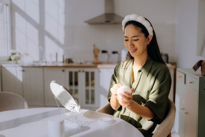 A woman is sitting at a table in her home, holding a skincare product. She looks happy while getting ready and is surrounded by a bright kitchen. Sunlight comes through the window.