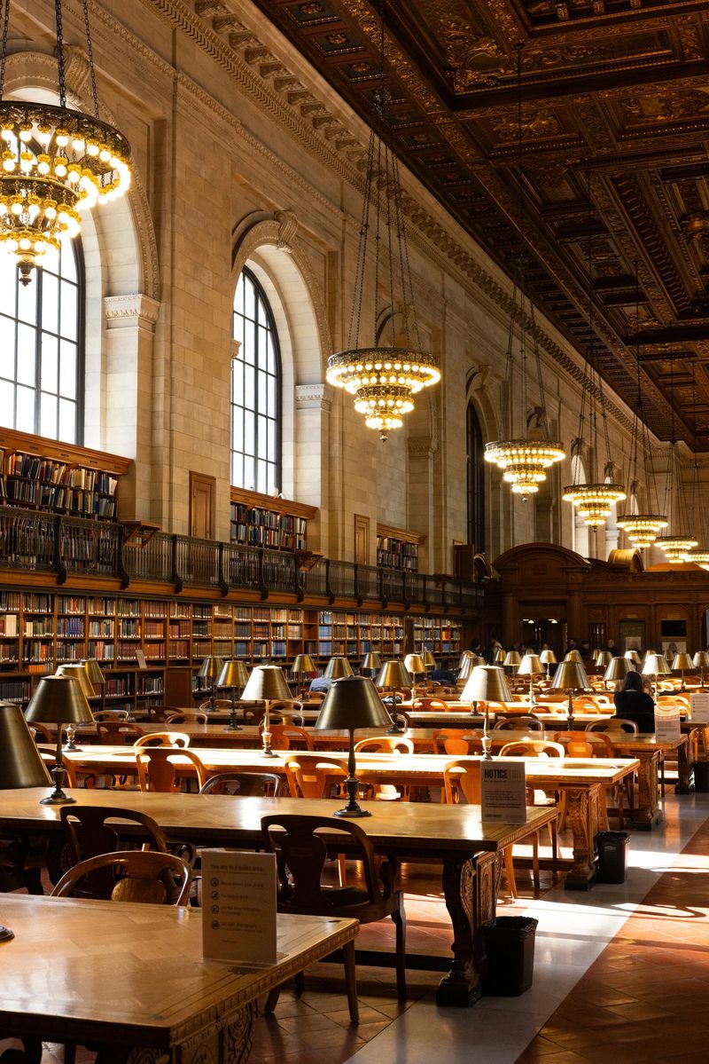 A grand library interior features rows of wooden tables with brass reading lamps, ornate chandeliers hanging from a detailed ceiling, large arched windows, and shelves filled with books, creating an elegant and quiet study environment.