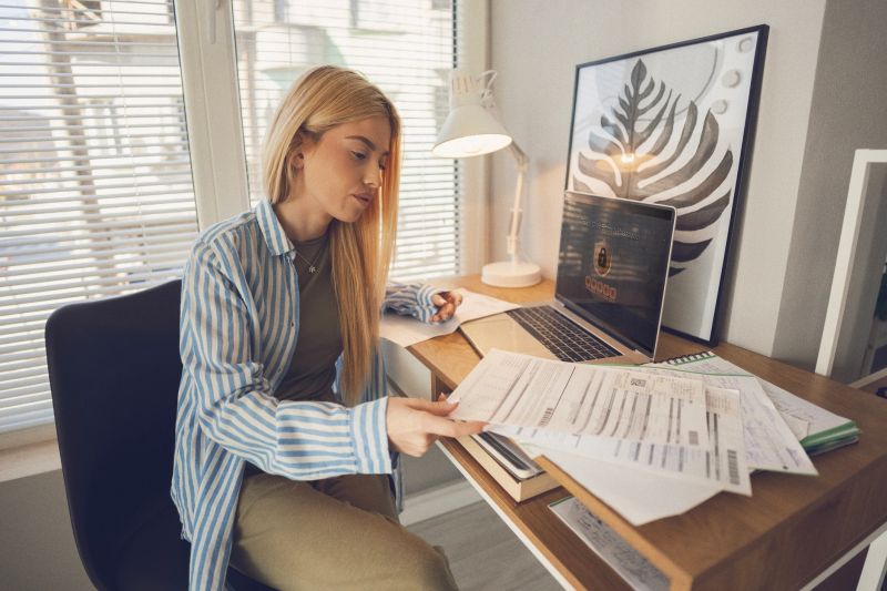 Young Gen Z business woman concentrating on paperwork management using network security with two-factor authentication on laptop screen, managing finances or contracts in her modern home office