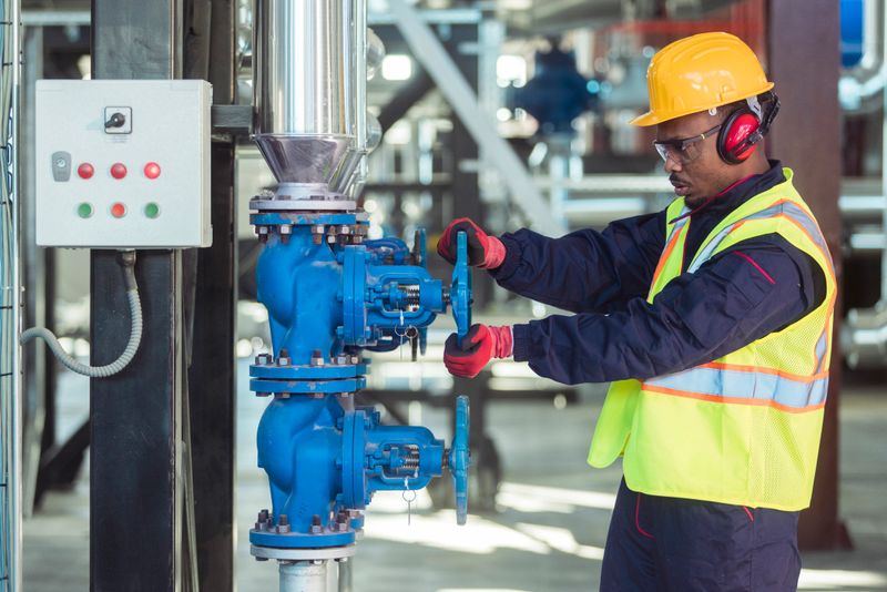 Male engineer wearing safety equipment checking and adjusting valve flow on industrial pipeline in processing plant