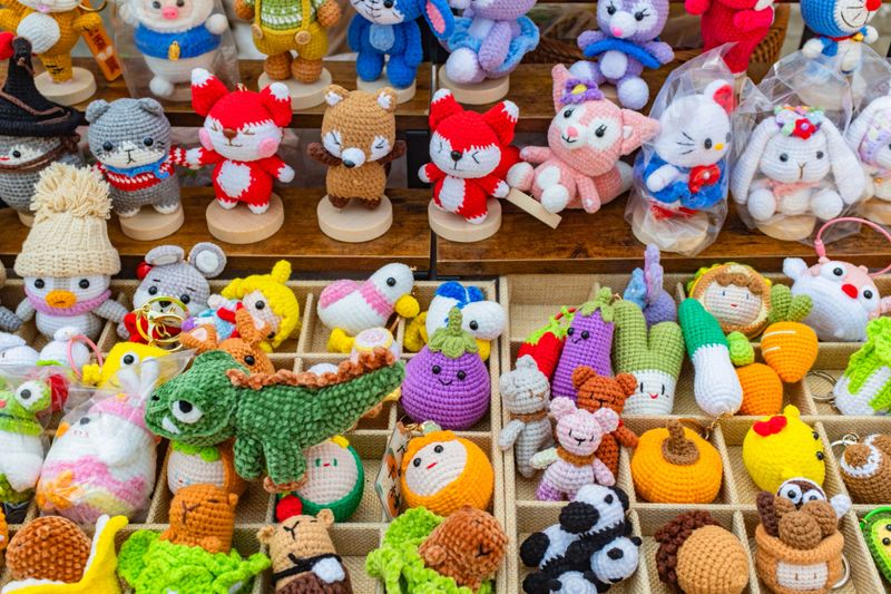 The image showcases a colorful and diverse collection of handmade crocheted plush toys, commonly known as amigurumi, neatly arranged for sale. The upper section features standing animal figures, including red foxes, cats, bunnies, and other characters, securely mounted on small round wooden bases. Below, divided wooden trays organize an assortment of smaller plush keychains and trinkets. This section prominently features a large green dinosaur, alongside cute anthropomorphic vegetables and fruits like a smiling purple eggplant, orange carrots, and a pumpkin. The compartments also hold tiny bears, pandas, acorns, and small character heads. The toys are crafted with bright, vibrant yarns, precise stitching, and endearing facial features, highlighting a charming, artisanal aesthetic typical of a boutique stall or craft fair.