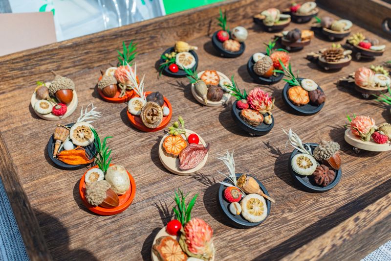 The photograph displays a shallow, textured wooden tray filled with neatly arranged, handmade botanical crafts presented under bright, direct sunlight. Each small, circular piece is built upon a flat base made of natural wood, painted black, or painted a vibrant orange. The miniature arrangements feature a charming, rustic assortment of natural, dried elements. These meticulously placed decorations include whole brown acorns, textured seed pods, dehydrated citrus slices, dried pink and yellow flower buds, and vibrant red berries, all accented with small sprigs of green foliage. The sharp, distinct shadows cast by the bright outdoor lighting emphasize the rich textures, three-dimensional depth, and earthy, autumnal color palette of these artisanal creations, capturing the lively aesthetic of an outdoor craft market display.