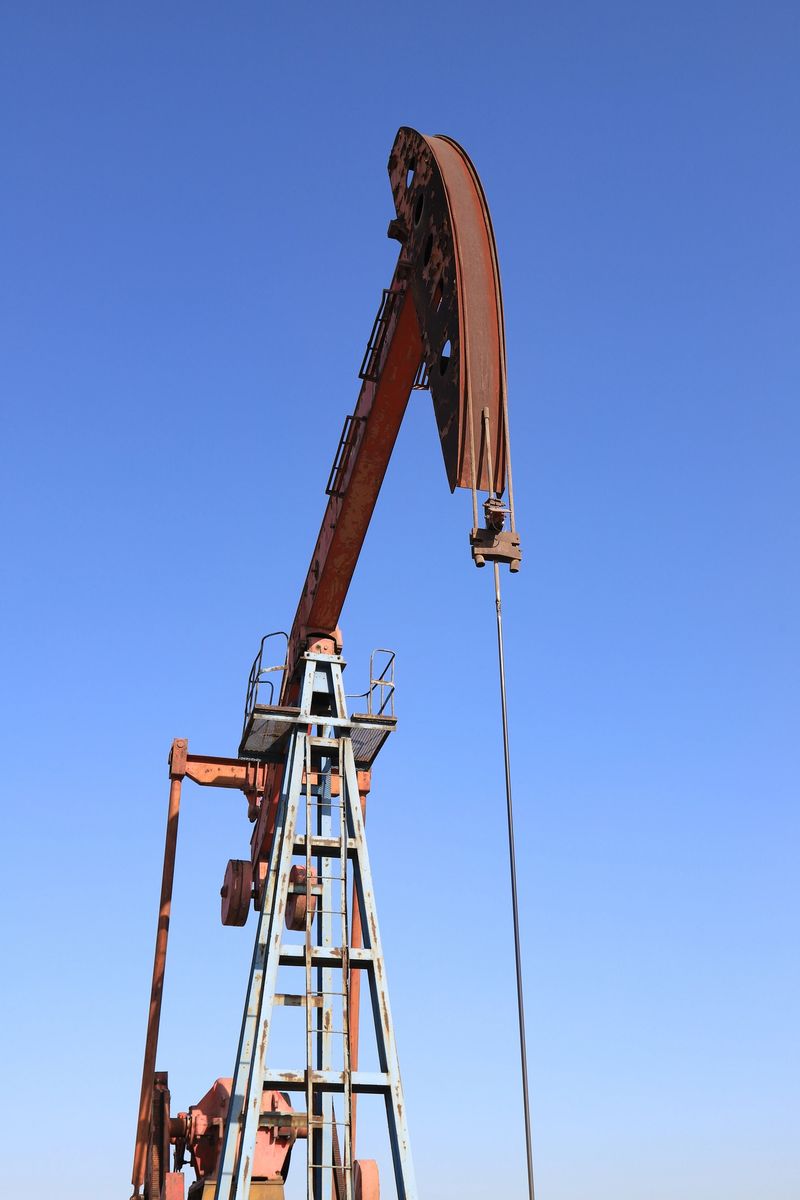 Close-up low-angle view of a weathered, rust-stained red oil pump jack (nodding donkey) in an oilfield. The industrial machinery stands prominently against a vivid, cloudless cerulean blue sky, showcasing the heavy industry of petroleum extraction and energy production.