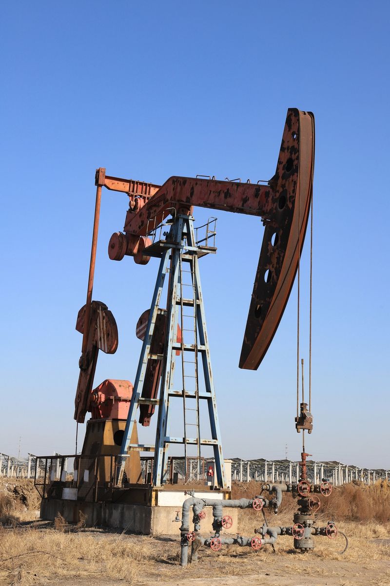 High-resolution photograph capturing an industrial oilfield scene under a bright, cloudless blue sky. The central subject is a large, weathered red oil pump jackcommonly known as a nodding donkeyin active operation. Below the machinery, connected oil production valves and pipelines are clearly visible, facilitating crude oil extraction. The background features dry wasteland vegetation and distant industrial infrastructure, embodying the raw, rugged essence of petroleum energy production and the heavy industry of resource extraction.