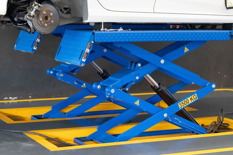 Blue car lift raises white vehicle. Heavy-duty lift marked 3500 KG capacity. Yellow safety markings on asphalt. Industrial garage setting. Ideal for automotive repair or maintenance scenes