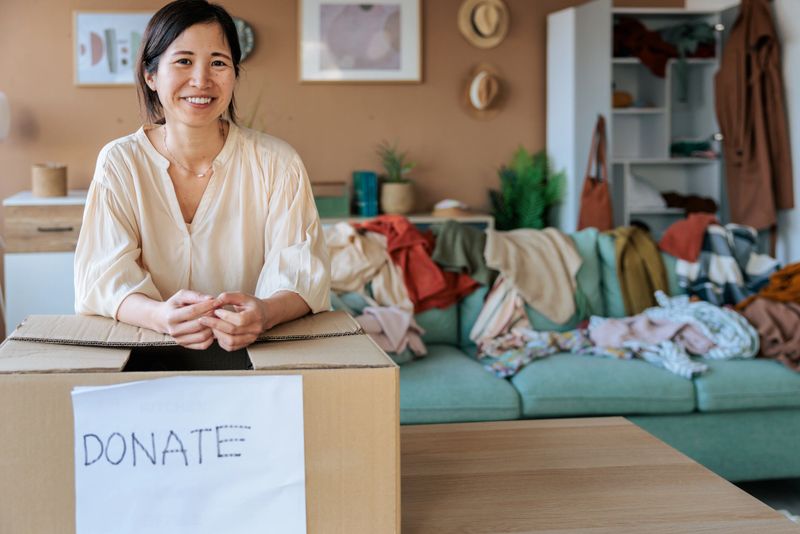 Smiling woman standing with a donation box after sorting and decluttering her clothes. The scene highlights positive emotions, organization, and giving away unused items.