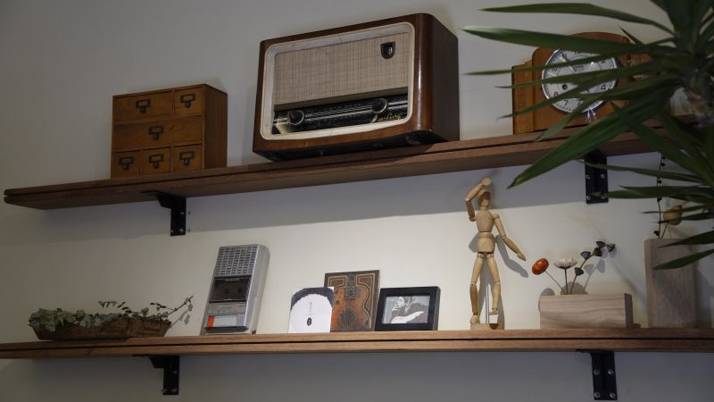 Retro wooden shelves display vintage radio, analog clock, wooden mannequin, cassette player, framed photos, and green plants in a cozy home decor setting with warm lighting.