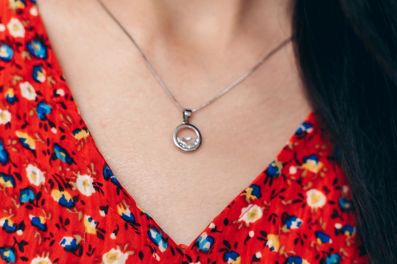 Macro shot of an elegant silver necklace with a round crystal pendant. The jewelry is highlighted on the neckline with a vibrant red floral dress, emphasizing detail, elegance, and feminine fashion style.