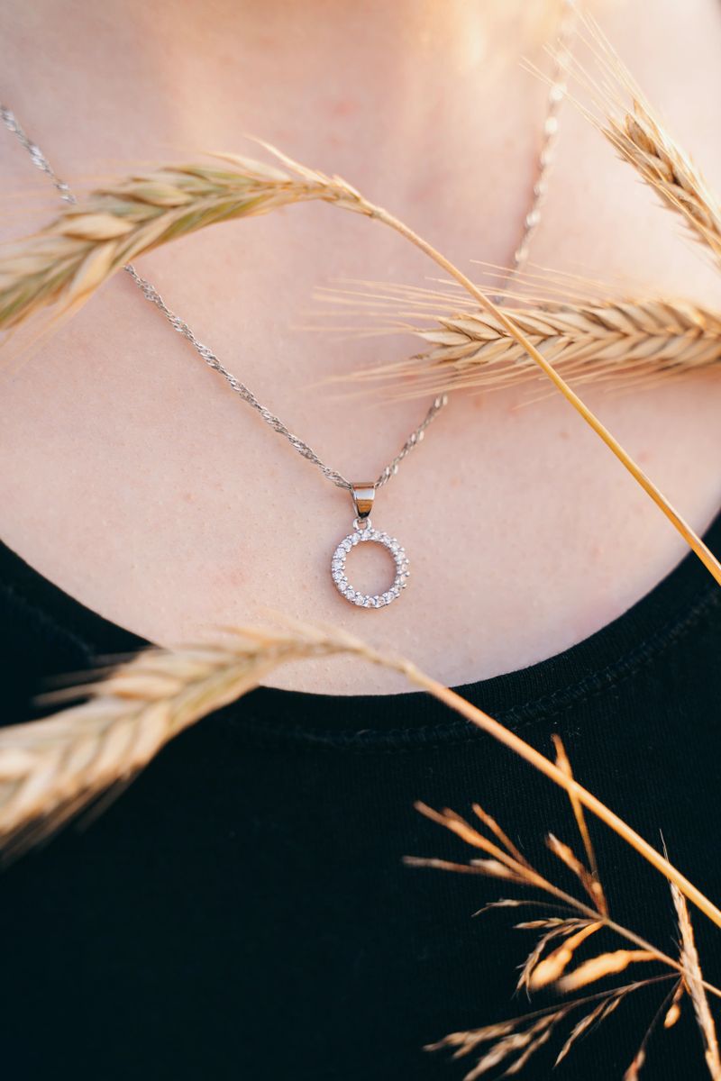 Close-up of a delicate silver necklace with a circular crystal pendant resting on a woman's neck. Captured through golden wheat stalks in soft evening sunlight, highlighting the fine chain and sparkle of the pendant. Ideal for jewelry promotions and editorial use.