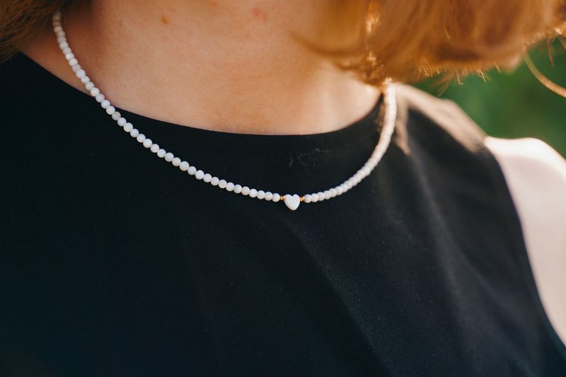 Detailed close-up of a delicate white beaded necklace with a small heart-shaped pendant, worn over a sleeveless black dress. The image highlights the texture and design of the jewelry in soft natural light, ideal for fashion, accessories, and jewelry-themed content.