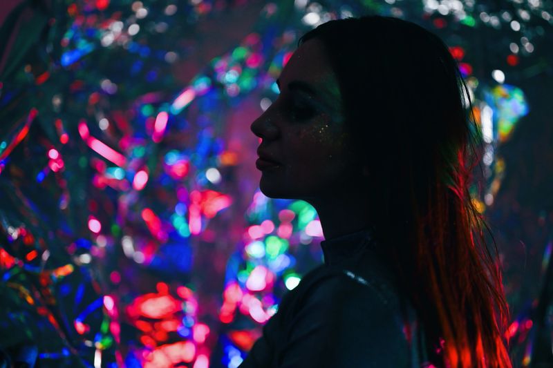 Woman standing in profile, lit by colorful bokeh lights and wearing face glitter