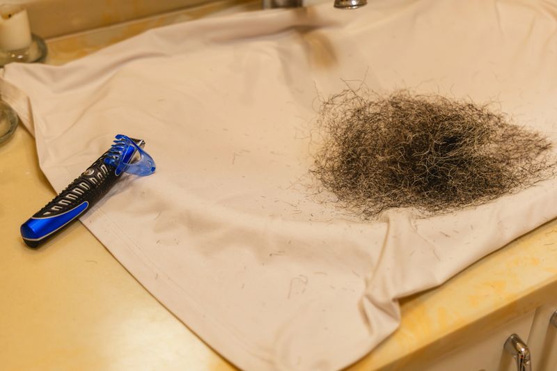 A white cloth lies on a bathroom sink. An electric razor rests beside a large pile of dark hair clippings. The scene shows a recent grooming session.