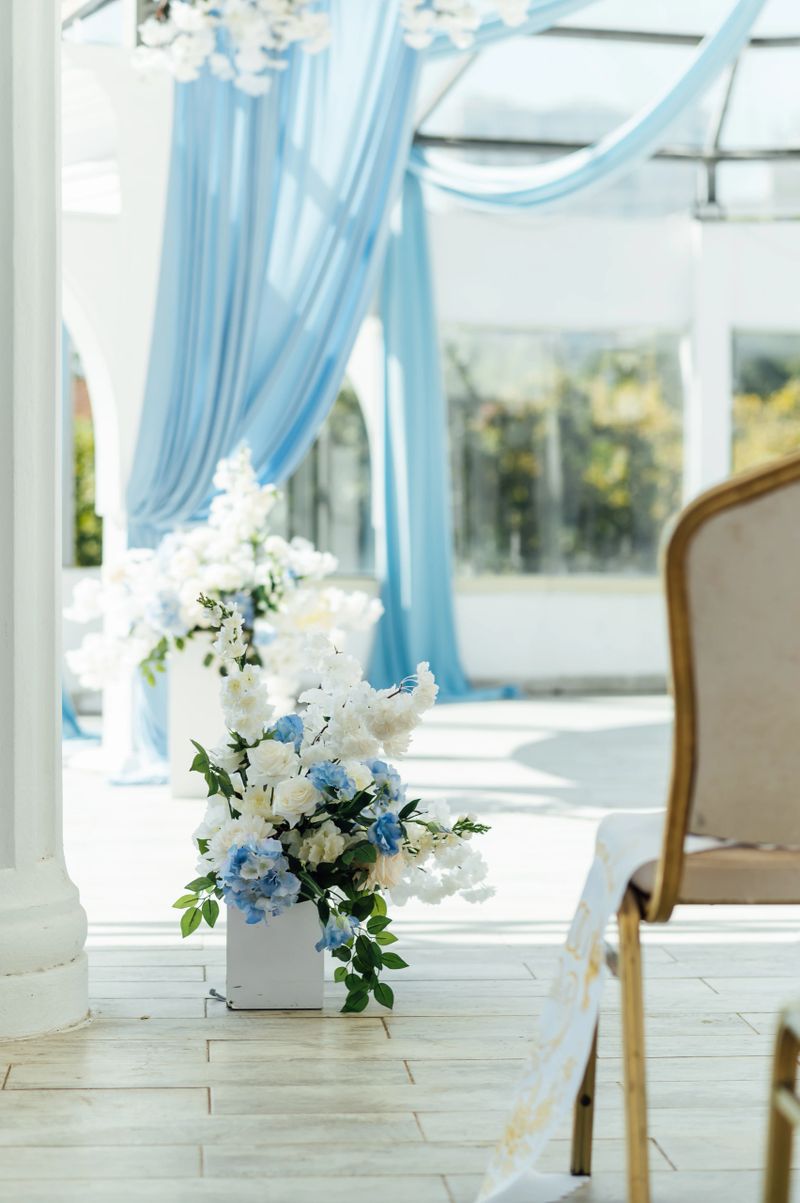 A collection of professional photographs showcasing a sophisticated and airy wedding ceremony setup. The series captures details and wide shots of a modern gazebo adorned with flowing sky blue curtains and lush arrangements of white and light-blue flowers. Key elements include tall white pedestals with large white floral centerpieces, light-blue drapes, delicate flower balls, a white aisle runner, and elegant guest chairs, all set under bright daylight on a spacious terrace.