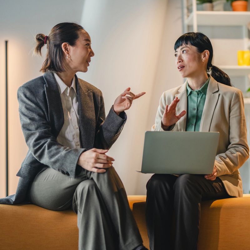 Two asian business women collaborating on a project, one actively explaining points while holding a laptop, the other listening intently, fostering engagement and teamwork in a modern office