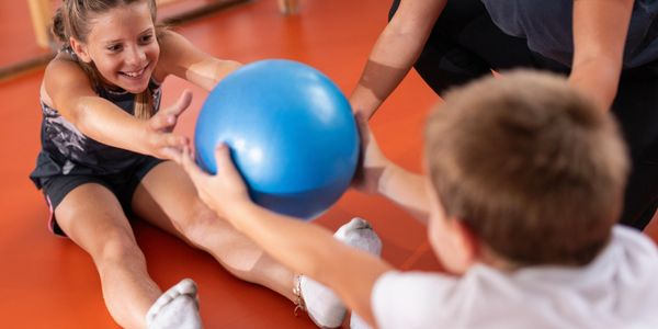 Two children passing a blue exercise ball while sitting on the floor.
