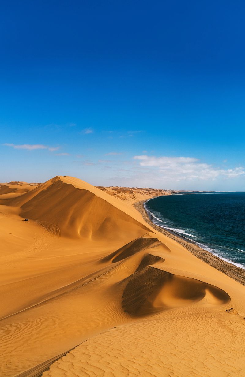 Golden sand dunes cascade into the deep blue ocean at Sandwich Bay near Swakopmund, Namibia, creating a dramatic coastal desert landscape that evokes solitude, adventure, and vast natural beauty.