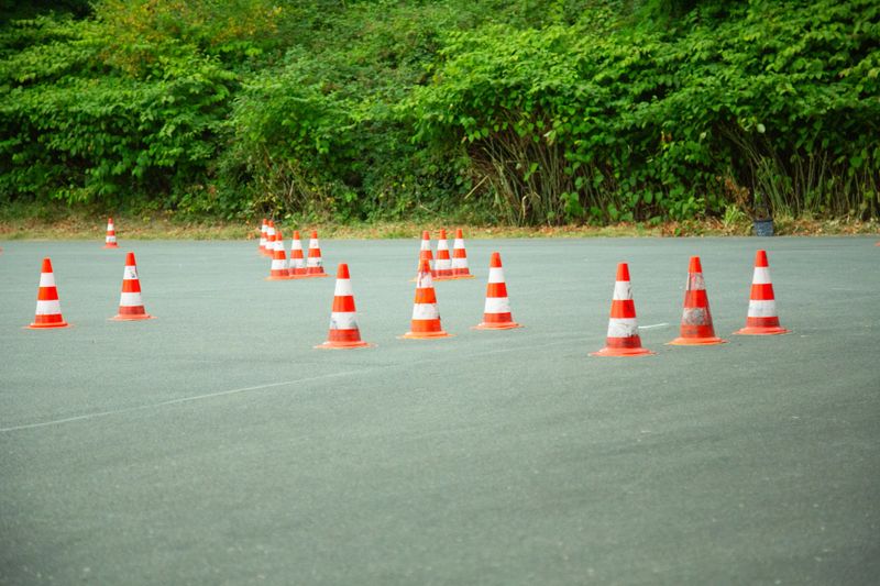 A wide asphalt platform is arranged with multiple bright orange traffic cones forming a structured driving practice course used for training and testing vehicle control skills. The cones create clear paths and obstacles that guide movement and simulate real road conditions in a safe environment. The surrounding greenery adds contrast and context to the scene while emphasizing that the location is outdoors. This composition highlights precision coordination and learning making it suitable for concepts related to driving school education transportation safety and professional training in controlled conditions.