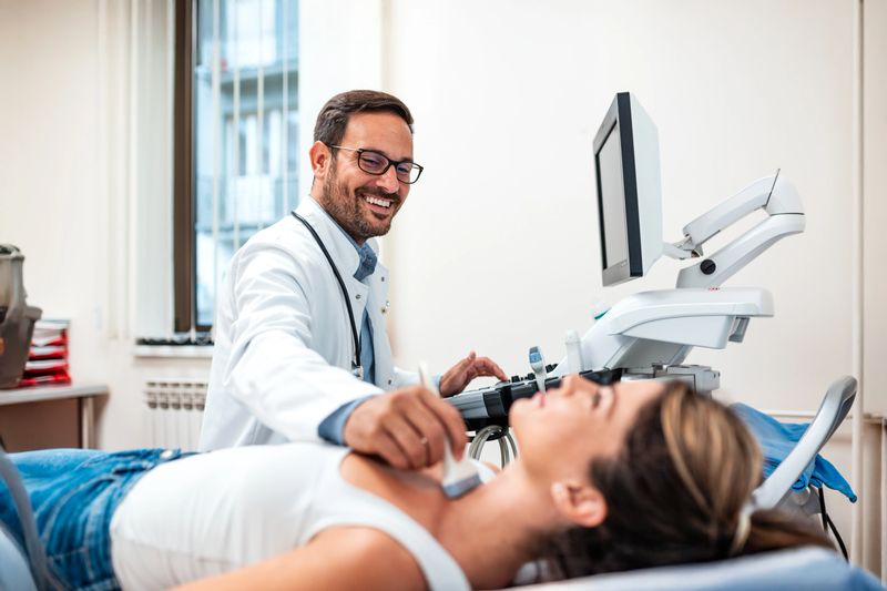 Smiling doctor performing ultrasound on female patient in modern clinic. Healthcare professional using sonogram for diagnosis. Medical imaging, wellness, and healthcare concept in hospital setting.