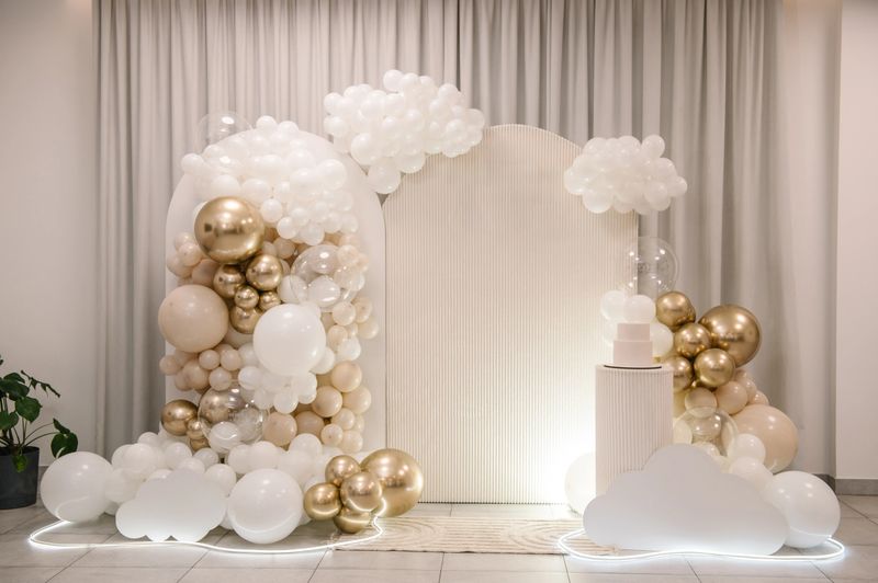 Arch. Elegant photo zone for a wedding or anniversary featuring a fluted backdrop, a white two-tier cake on a pedestal, and a luxurious organic balloon arch in white, beige, gold colors in restaurant.