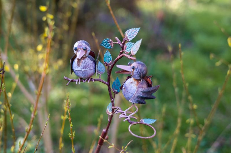Two metal bird sculptures perch on a decorative branch with blue leaves, adding charm to the garden. Their presence brings a whimsical touch to the outdoor space.