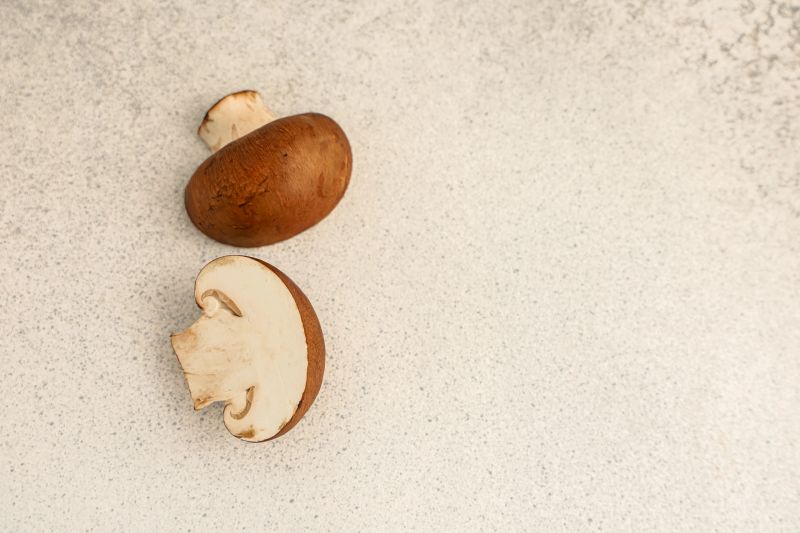 Mushrooms are placed on a light surface in a kitchen. One mushroom is whole while the other is sliced in half. The setting is bright and inviting.