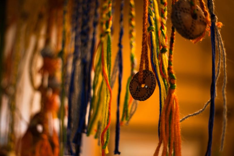 A vibrant close-up of handmade ethnic decorations. The image features multi-colored braided threads and strings adorned with circular clay pendants. The warm, soft bokeh background emphasizes the intricate textures of the traditional folk craft.