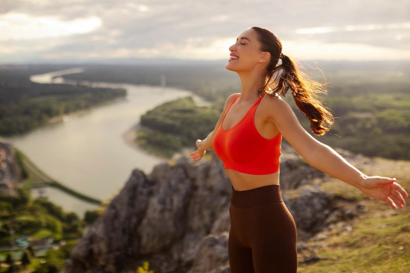 Woman doing yoga on mountain overlooking river, lady spreading hands and breathing, enjoying fresh air and outdoors workout
