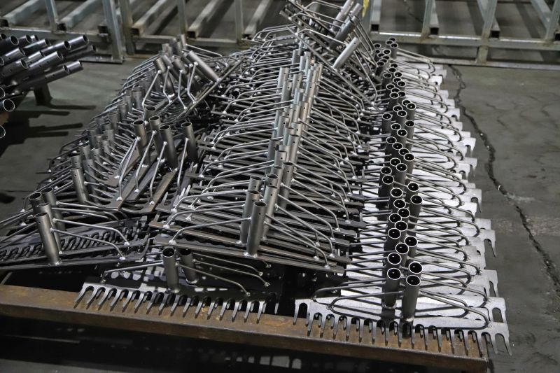 The image captures the interior of an industrial production workshop, where a large number of metal rake blanks are neatly and densely stacked on industrial brackets. The components feature a uniform U-shaped hook and long handle structure, with black oxidized surfaces. It clearly presents the industrial production scene of hardware farm tool processing and mass preparation, suitable for material use in fields such as farm tool manufacturing and metal processing.