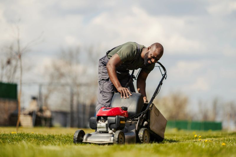 Mid-40s man in overalls prepping a push mower in a sunny green garden, smiling as he performs routine lawn maintenance and outdoor spring/summer yard care