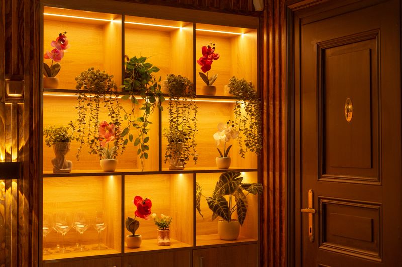 Warm wooden shelf with orchids backlit by amber lighting, potted plants, glass vases, small decorative sculptures. Interior details
