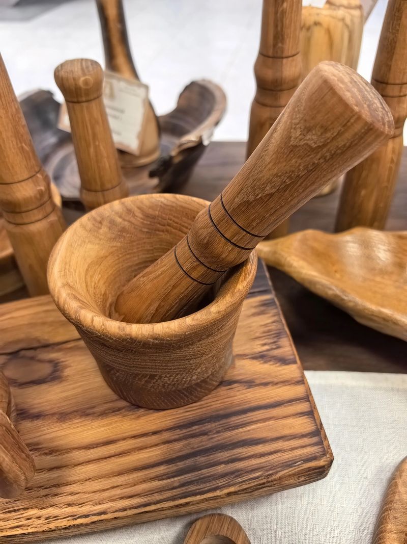A beautifully crafted wooden mortar and pestle sits prominently on display. Surrounded by various handcrafted wooden items, the scene captures the essence of artisan craftsmanship in a lively market.