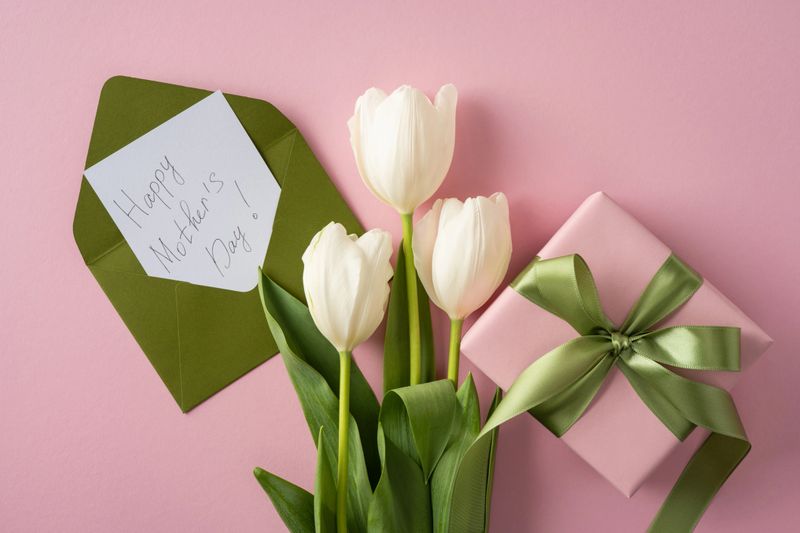 Happy Mother's Day arrangement with white tulips, a handwritten card in a green envelope and a pink gift box tied with satin green ribbon evoking spring love and thoughtful celebration