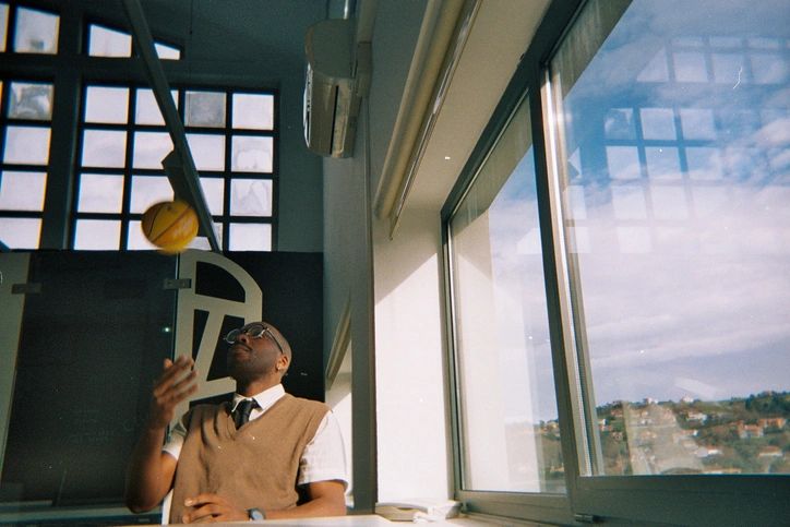 Young adult man playing with a small basketball indoors, wearing glasses and a sweater vest, showing a moment of reflection and leisure