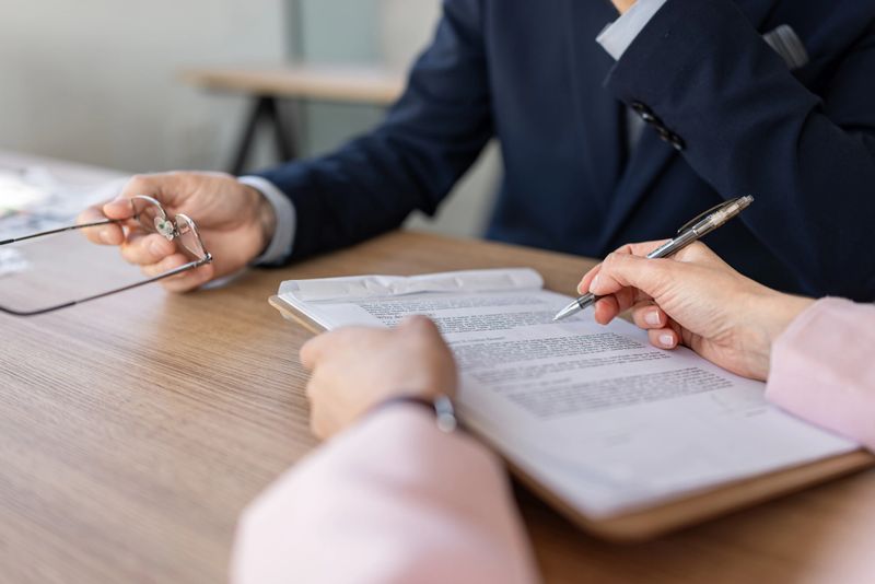 Business professionals review and sign contract documents at a wooden desk during a consultation, with a pen pointing to text and hands holding glasses, conveying trust and agreement.