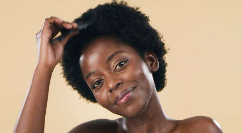 Confident Black woman gently combs her natural afro in a warm studio portrait, smiling with radiant skin and relaxed expression to convey self-care, natural beauty, confidence, and healthy hair.