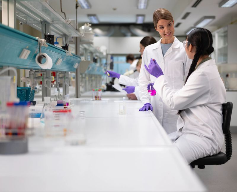 Caucasian female professor wearing white lab coat and purple gloves instructs diverse lab students at laboratory workbench. Students work with test tubes and samples under bright fluorescent lighting in professional research facility.