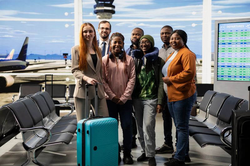 Portrait of diverse travelers waiting at departures lounge in busy airport terminal, checking flight information on screen and their boarding pass. Men, women and business passengers with luggage.
