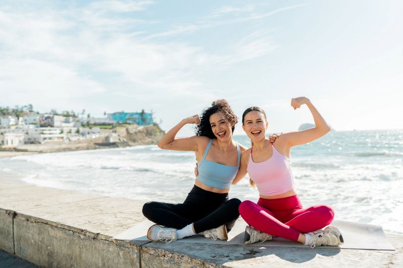 Portrait of young friends sportswomen flexing muscles and celebrating on sidewalk beach