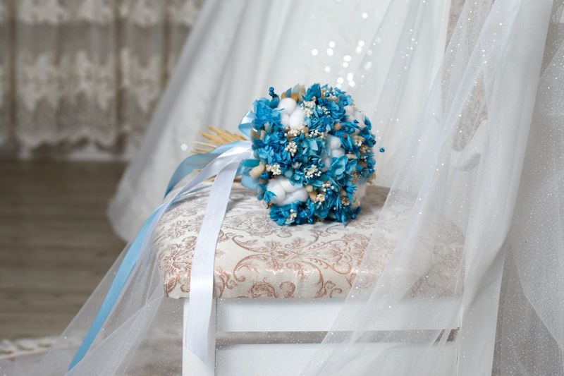 A bridal bouquet made of blue flowers is set on a chair in a wedding venue. Sunlight shines through the windows, creating a soft glow around the decorations.