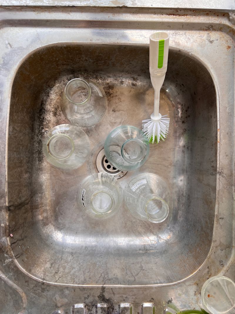 Overhead view of a metal sink with glass jars and a bottle brush, five clear glass jars arranged around drain, worn stainless steel surface, cleaning scene