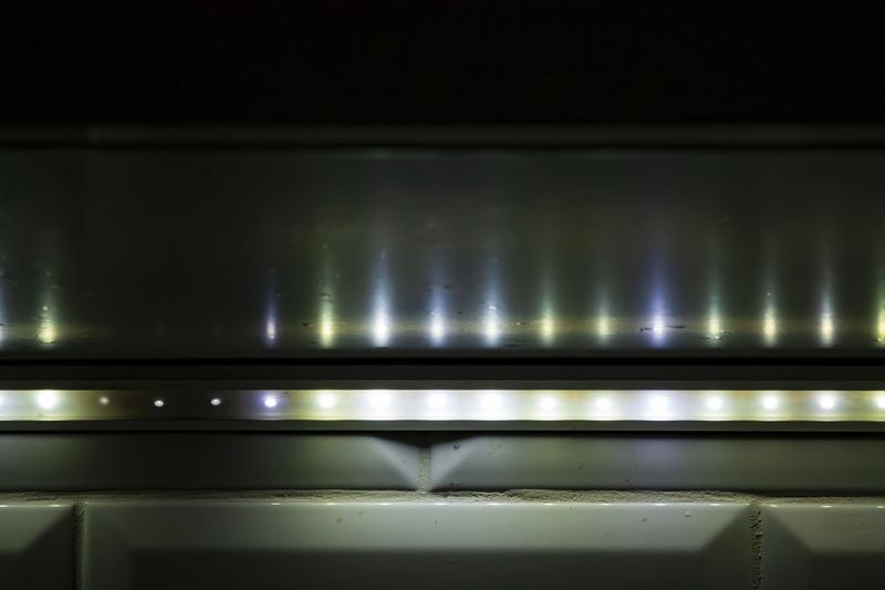 Close-up of under-cabinet led strip lights casting reflections on a glossy surface above and illuminating a tiled backsplash in dim setting