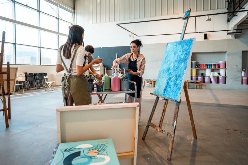 A diverse group of artists, including women and men, collaborates in a spacious, well-lit art studio filled with colorful supplies, easels, and large windows. They are painting and expressing creativity in a vibrant environment.