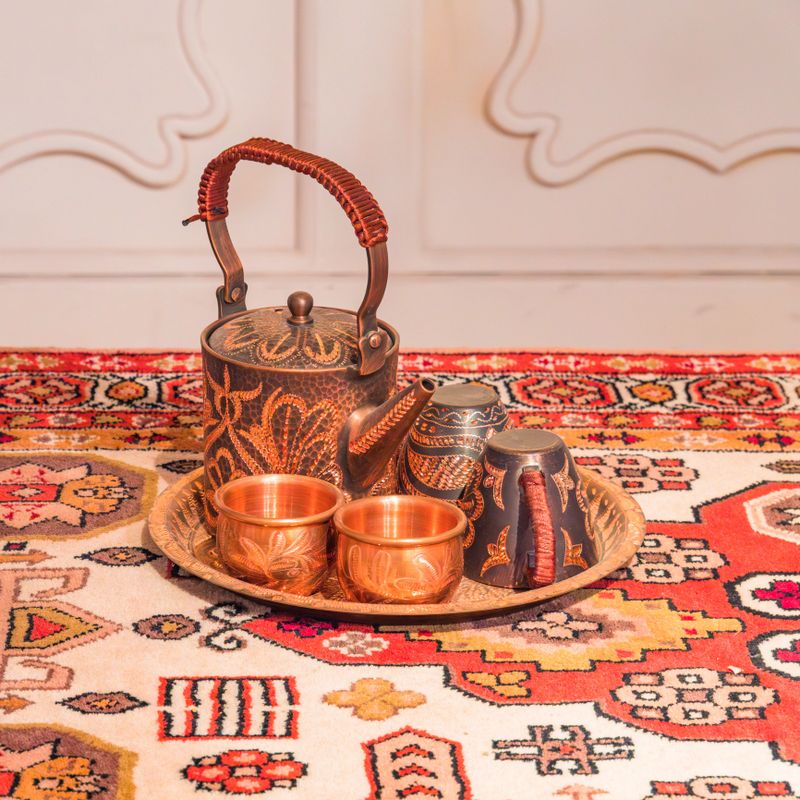 The photograph captures a beautifully crafted, artisanal copper tea set resting on a shallow, circular tray. The centerpiece is a stout teapot featuring a dark, textured patina beautifully contrasted by bright, deeply engraved copper floral and leaf motifs. Its high, arched metal handle is tightly wrapped in a reddish-brown woven cord. Accompanying the teapot are four cups in two distinct styles: two are gleaming, highly polished copper with delicate engravings, while the other two match the dark, oxidized finish of the teapot and feature similarly wrapped handles. The entire set is displayed on a vibrant, intricately woven traditional rug adorned with bold geometric patterns in shades of bright red, cream, gold, and black. A softly blurred, light-colored paneled wall in the background allows the warm metallic tones and rich colors of the foreground to stand out prominently under bright, warm lighting.