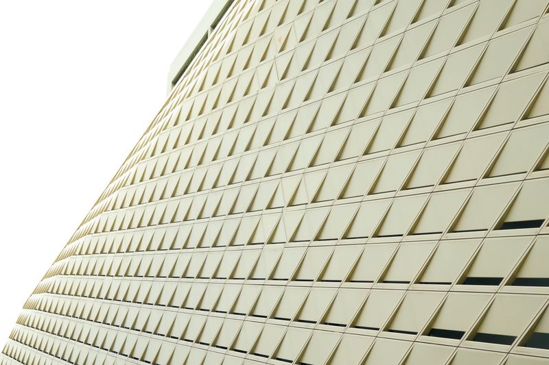Low-angle shot capturing the intricate geometric facade of a modern high-rise building. The surface features a repeating pattern of light beige diamond-shaped panels, creating a sense of depth and futuristic design. Lush green trees peek from the corner, blending nature with contemporary architecture.